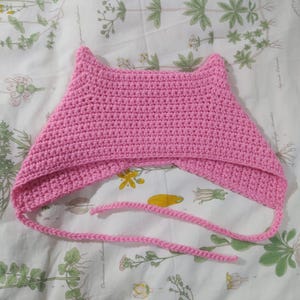 以下が含まれることがあります： ピンクの毛糸で編まれた猫耳フードで、長い紐が付いています。フードはシングルクロッシェで編まれており、シンプルなデザインです。