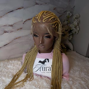 Puede incluir: Una peluca rubia trenzada sobre una cabeza de maniquí. La peluca presenta una parte delantera de encaje y largas trenzas sueltas. El maniquí lleva una camiseta rosa de manga larga con el texto "Aura LUXE HAIR" en la parte delantera. El fondo es una pared de mármol.