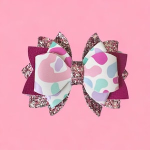 May include: A pink and white patterned hair bow with a pink glitter trim. The bow is made of fabric and has a white background with pink, purple, and blue abstract shapes.