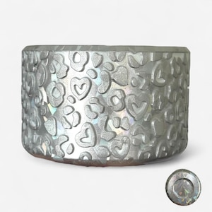 May include: A silver-colored, cylindrical container with a textured surface featuring a pattern of hearts and leopard spots. The container has a matching lid. The design is embossed on the surface, creating a raised effect.