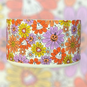 May include: A white ceramic candle holder with a Halloween-themed design. The design features a pattern of purple, orange, and yellow flowers with ghosts, pumpkins, and candy corn accents.