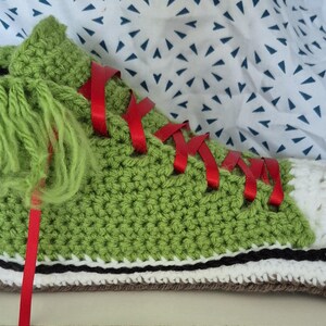 May include: A handmade, green crochet trainer with a white toe, red ribbon laces, and a shaggy green tongue. The shoe has a black and white stripe along the sole and a brown sole. The trainer is a unique, whimsical design.