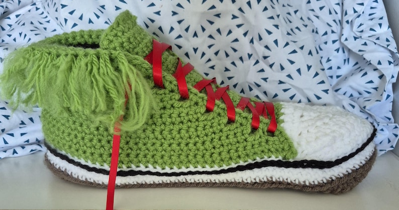 May include: A handmade, green crochet trainer with a white toe, red ribbon laces, and a shaggy green tongue. The shoe has a black and white stripe along the sole and a brown sole. The trainer is a unique, whimsical design.
