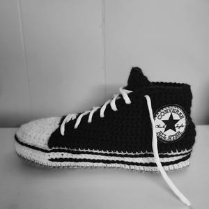 May include: A black and white crocheted high-top sneaker with white laces. The sneaker features a white toe cap and a white sole with black stripes. A patch on the side reads "CONVERSE Chuck Taylor ALL STAR".