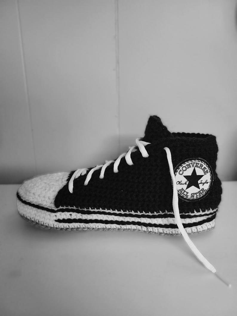 May include: A black and white crocheted high-top sneaker with white laces. The sneaker features a white toe cap and a white sole with black stripes. A patch on the side reads "CONVERSE Chuck Taylor ALL STAR".