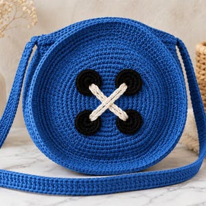 Peut inclure: Un sac bandoulière rond en crochet bleu vif, orné d'un grand motif de bouton décoratif sur le devant. Le bouton est composé de quatre petits cercles noirs reliés par deux épais fils de laine couleur crème formant une croix.