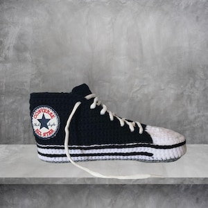 May include: A black and white crochet high-top sneaker with white laces. The sneaker features the Converse All Star logo on the side. The toe and sole are white, with two white stripes on the midsole. The sneaker is displayed on a gray surface.