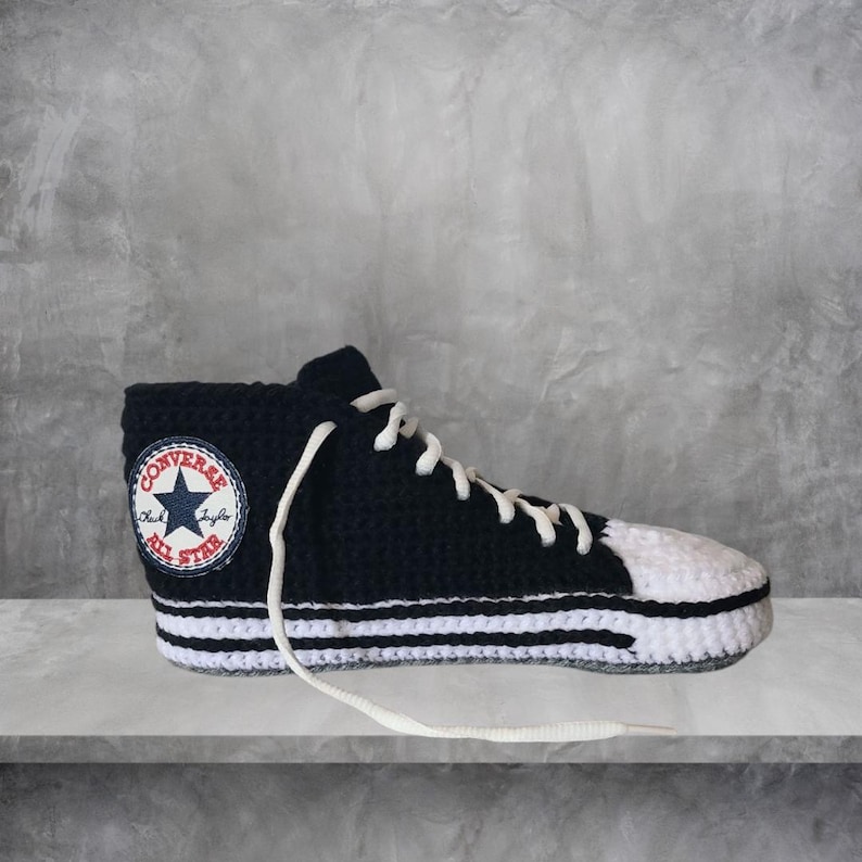 May include: A black and white crochet high-top sneaker with white laces. The sneaker features the Converse All Star logo on the side. The toe and sole are white, with two white stripes on the midsole. The sneaker is displayed on a gray surface.