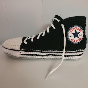Peut inclure: Une basket montante noire et blanche au crochet avec des lacets blancs. La basket a un bout blanc et un écusson circulaire avec les mots "Converse All Star" et une étoile bleue. La chaussure a des rayures blanches autour de la semelle.
