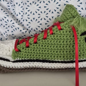 May include: A handmade, crocheted trainer in shades of green, white, and brown. The shoe features red laces and a black and yellow graphic on the side. The shoe is designed to resemble a cartoon character.