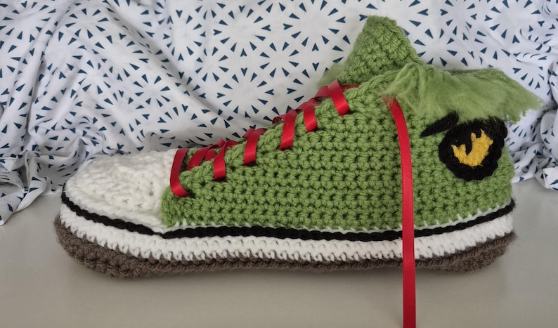 May include: A handmade, crocheted trainer in shades of green, white, and brown. The shoe features red laces and a black and yellow graphic on the side. The shoe is designed to resemble a cartoon character.