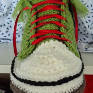 May include: A crocheted shoe in green, white, black, and brown. It has red ribbon laces and a white toe cap. The shoe is a unique, handcrafted item, likely for decorative purposes.