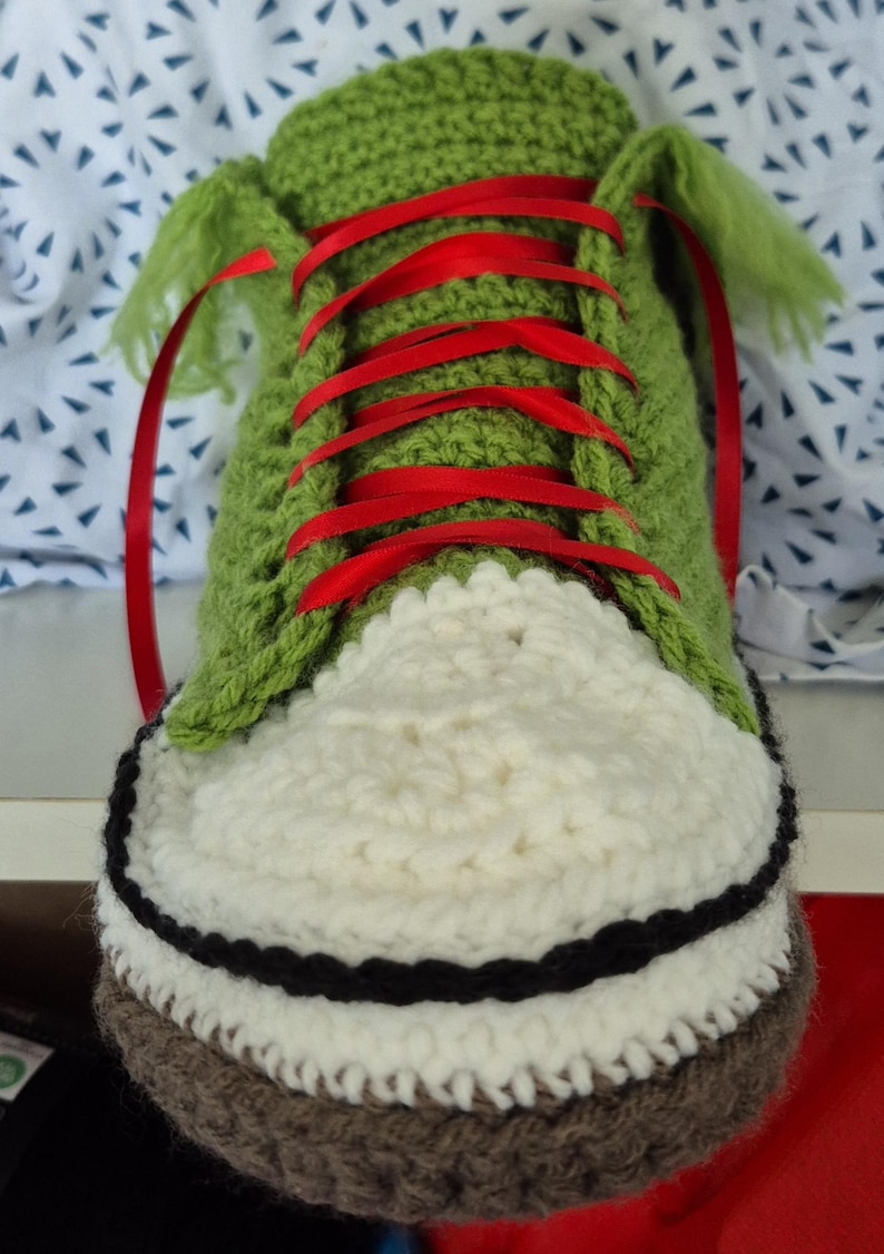 May include: A crocheted shoe in green, white, black, and brown. It has red ribbon laces and a white toe cap. The shoe is a unique, handcrafted item, likely for decorative purposes.