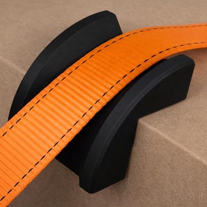 May include: Close-up of a black object holding an orange strap. The strap has black stitching and a textured surface. The black object is curved and appears to be made of plastic or rubber.