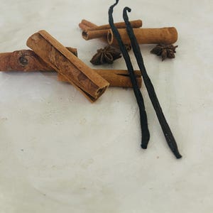 K&ouml;nnte beinhalten: Zwei lange, dunkelbraune Vanilleschoten und mehrere Zimtstangen mit Sternanis auf einer wei&szlig;en Oberfl&auml;che.
