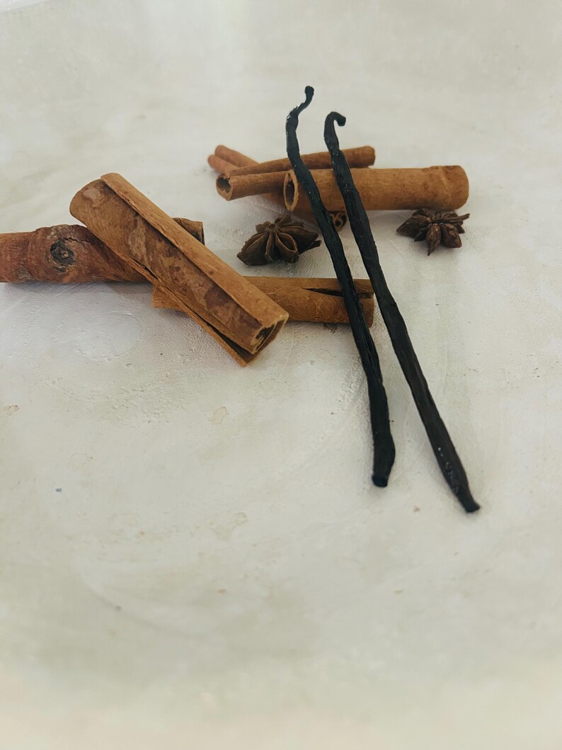 K&ouml;nnte beinhalten: Zwei lange, dunkelbraune Vanilleschoten und mehrere Zimtstangen mit Sternanis auf einer wei&szlig;en Oberfl&auml;che.