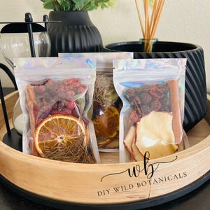 K&ouml;nnte beinhalten: Vier durchsichtige Plastikt&uuml;ten gef&uuml;llt mit getrockneten Fr&uuml;chten, Gew&uuml;rzen und Kr&auml;utern. Die Beutel sind mit "DIY Wild Botanicals" beschriftet und auf einem Holztablett angeordnet.