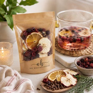 May include: A brown paper pouch with a clear window containing dried fruit slices, berries, and spices, labeled with the text "wb DIY WILD BOTANICALS". The pouch sits next to a glass teapot filled with a fruit infusion and a small wooden plate of ingredients.
