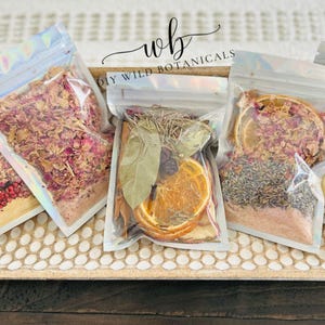 May include: Clear, resealable bags filled with dried botanicals, including rose petals, orange slices, and herbs. The bags are arranged on a decorative tray. The text "wb DIY WILD BOTANICALS" is visible in the image.