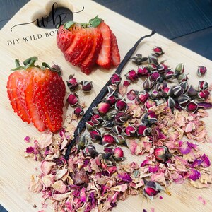 Puede incluir: Una tabla de cortar de madera con una vaina de vainilla, fresas frescas y pétalos de rosa secos. El texto "DIY WILD BOTANICALS" está escrito en la tabla.