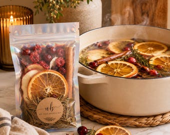 Op de afbeelding: Een doorzichtige, hersluitbare zak met gedroogde sinaasappelschijfjes, rode veenbessen, een kaneelstokje en rozemarijntakjes, met een rond etiket "wb DIY WILD BOTANICALS". Daarnaast staat een grote, crèmekleurige pan gevuld met sudderende vloeistof.