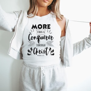 Könnte beinhalten: Weißes Tanktop mit dem Text "MORE than a Conqueror through Christ" in Schwarz. Das Tanktop wird mit einem grauen Kapuzenpullover mit Reißverschluss und einer grauen Jogginghose getragen. Der Text ist in einer fetten, stilisierten Schriftart.