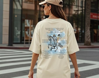 May include: Cream-colored oversized t-shirt featuring a graphic of a figure on horseback with wings, accompanied by text. The design is in shades of blue, white, and gray. The shirt is being worn by a person.