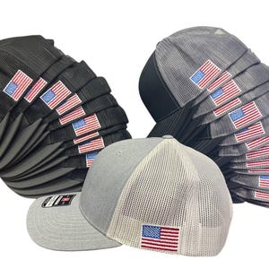 May include: A collection of baseball caps in various shades of gray and black, each featuring an embroidered American flag patch. The caps have a structured design with a mesh back for breathability.