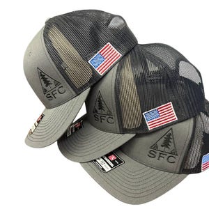May include: A stack of gray trucker hats with black mesh backs. Each hat features an embroidered American flag patch and a triangular logo with the letters "SFC". The hats are a neutral color.