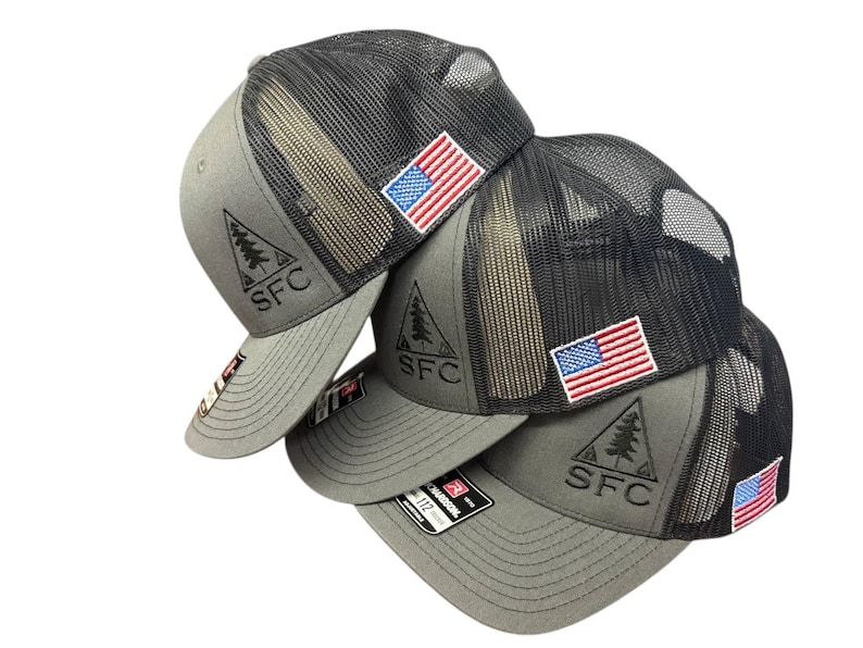 May include: A stack of gray trucker hats with black mesh backs. Each hat features an embroidered American flag patch and a triangular logo with the letters "SFC". The hats are a neutral color.