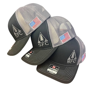 May include: Four black and gray trucker hats with the American flag patch and the letters "SFC" embroidered on the front. The hats have a mesh back and a curved brim. The hats are stacked on top of each other.
