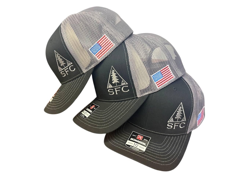 May include: Four black and gray trucker hats with the American flag patch and the letters "SFC" embroidered on the front. The hats have a mesh back and a curved brim. The hats are stacked on top of each other.