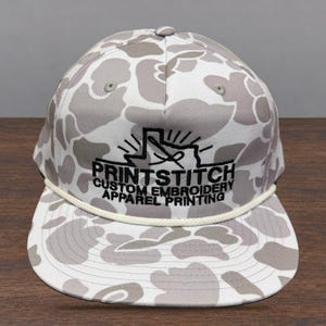 May include: A white and gray camouflage baseball cap featuring a white rope detail across the base of the crown. The front displays black embroidered text reading PRINTSTITCH CUSTOM EMBROIDERY APPAREL PRINTING with a Texas state outline graphic.