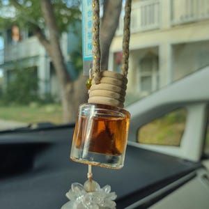 Pode incluir: Uma garrafa de vidro marrom com uma tampa de madeira pendurada no espelho retrovisor de um carro. A garrafa est&aacute; cheia de um l&iacute;quido transparente e tem um amuleto de flor branca pendurado abaixo. A garrafa tem uma etiqueta com caracteres chineses.