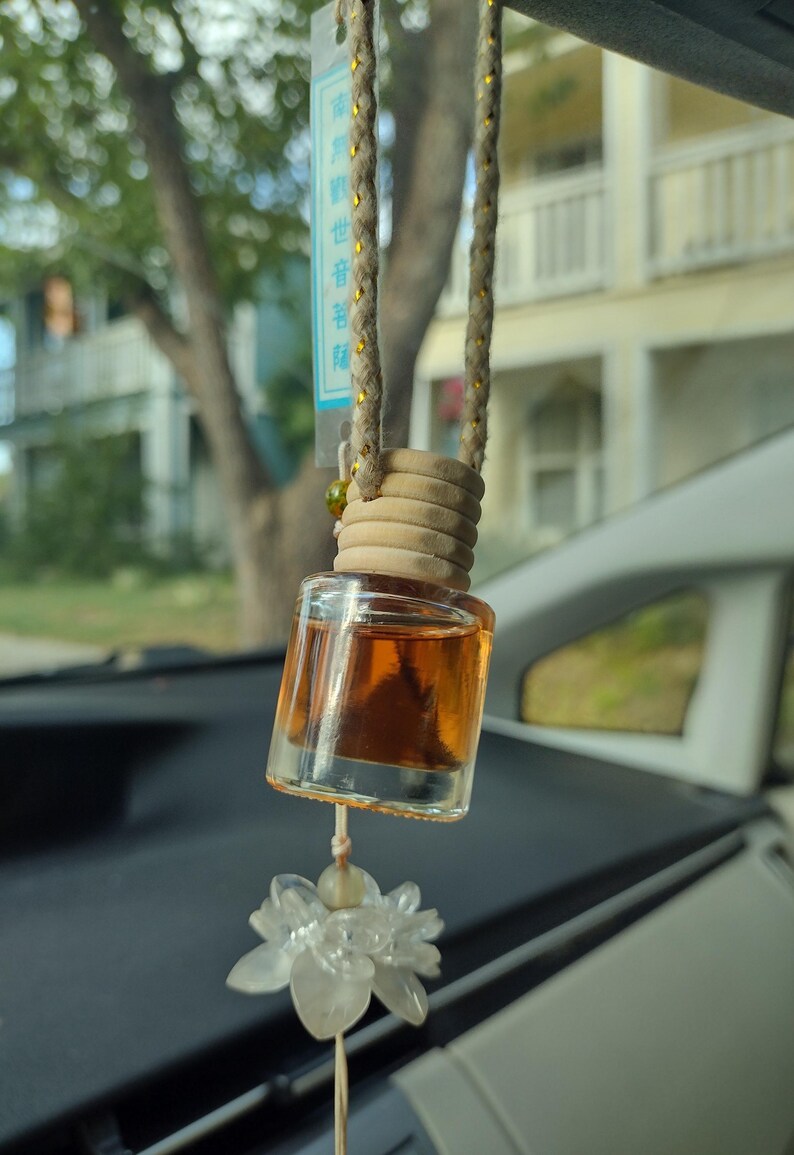 Pode incluir: Uma garrafa de vidro marrom com uma tampa de madeira pendurada no espelho retrovisor de um carro. A garrafa est&aacute; cheia de um l&iacute;quido transparente e tem um amuleto de flor branca pendurado abaixo. A garrafa tem uma etiqueta com caracteres chineses.