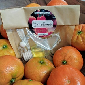 May include: A selection of fresh oranges surrounds an air freshener with a wooden diffuser. The label on the packaging reads "Bowl of Oranges" and "Flame and Florals". The air freshener contains approximately 8ml of liquid.