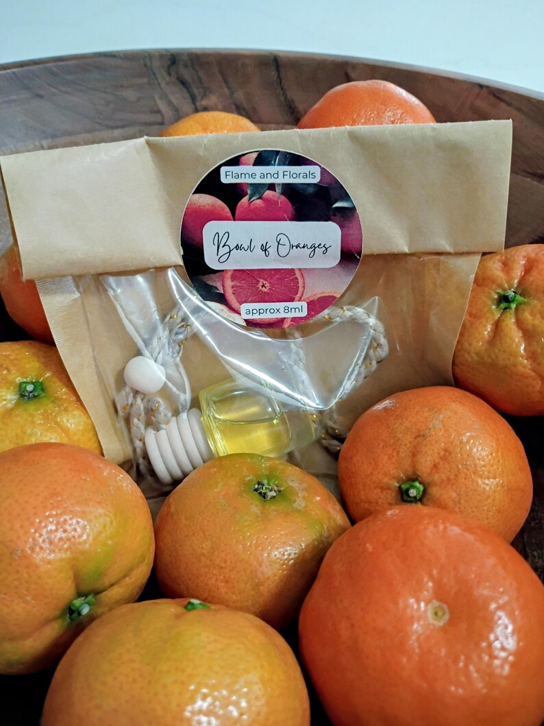 May include: A selection of fresh oranges surrounds an air freshener with a wooden diffuser. The label on the packaging reads "Bowl of Oranges" and "Flame and Florals". The air freshener contains approximately 8ml of liquid.