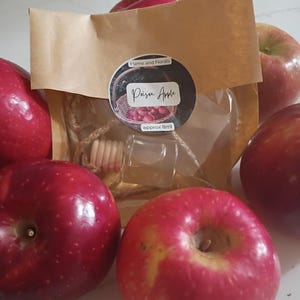 May include: A selection of red apples surrounds a small glass bottle with a wooden top, packaged in a brown paper bag. The bag has a circular sticker that reads "Flame and Florals" and "Poison Apple", with "approx 8ml" below. The apples are shiny and vibrant.