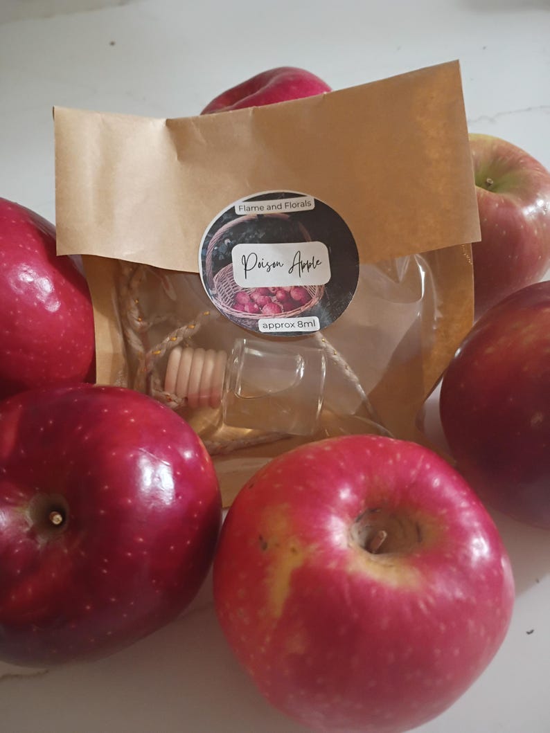May include: A selection of red apples surrounds a small glass bottle with a wooden top, packaged in a brown paper bag. The bag has a circular sticker that reads "Flame and Florals" and "Poison Apple", with "approx 8ml" below. The apples are shiny and vibrant.