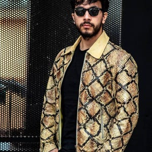 Peut inclure: Un homme portant une veste en cuir à motif de peau de serpent jaune et marron avec une chemise noire en dessous. Il porte des lunettes de soleil et a une barbe.