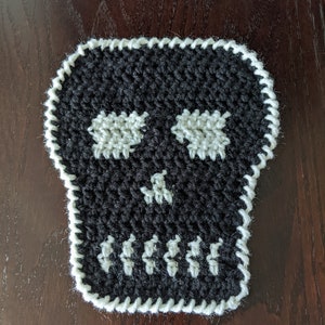 Könnte beinhalten: Ein schwarzer gehäkelter Schädel mit weißen gehäkelten Augen und Zähnen. Der Schädel hat einen weißen gehäkelten Rand.