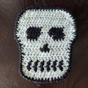 Könnte beinhalten: Ein weißer gehäkelter Schädel mit schwarzen Garn-Details. Der Schädel hat zwei Augenhöhlen, eine Nase und einen Mund.