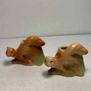 May include: Two ceramic squirrel figurines. The squirrels are in a sitting position with their tails up. The squirrels are tan, orange, and green. The figurines have a hole in the back.