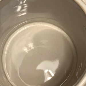 Peut inclure: Mug en c&eacute;ramique cr&egrave;me avec un int&eacute;rieur brillant. L'int&eacute;rieur de la tasse pr&eacute;sente des fissures visibles. Le bord et la anse de la tasse sont visibles, avec un motif textur&eacute; sur la anse.