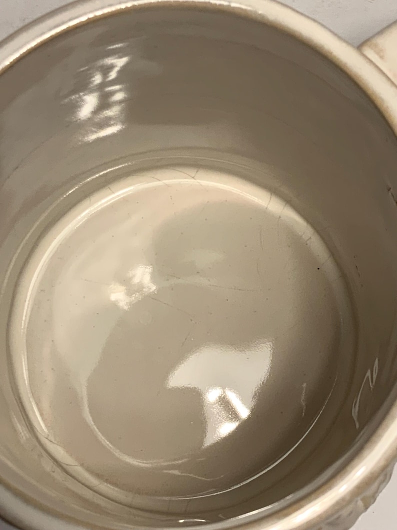 Peut inclure: Mug en c&eacute;ramique cr&egrave;me avec un int&eacute;rieur brillant. L'int&eacute;rieur de la tasse pr&eacute;sente des fissures visibles. Le bord et la anse de la tasse sont visibles, avec un motif textur&eacute; sur la anse.