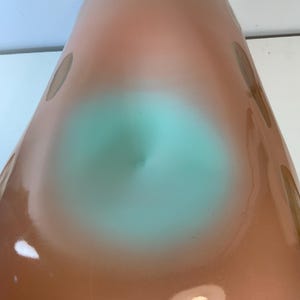 May include: A tall, cone-shaped vase with a gradient of colors. The top is a light peach, transitioning to a turquoise center, and then back to peach. Brownish-gray spots are scattered across the surface. The vase has a glossy finish.