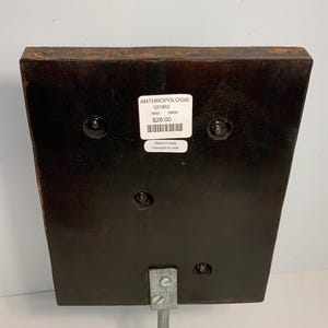May include: A dark brown wooden decorative piece with a metal stand. The piece has a rectangular shape with rounded edges and is attached to a silver metal stand. A white label with text is visible on the front.