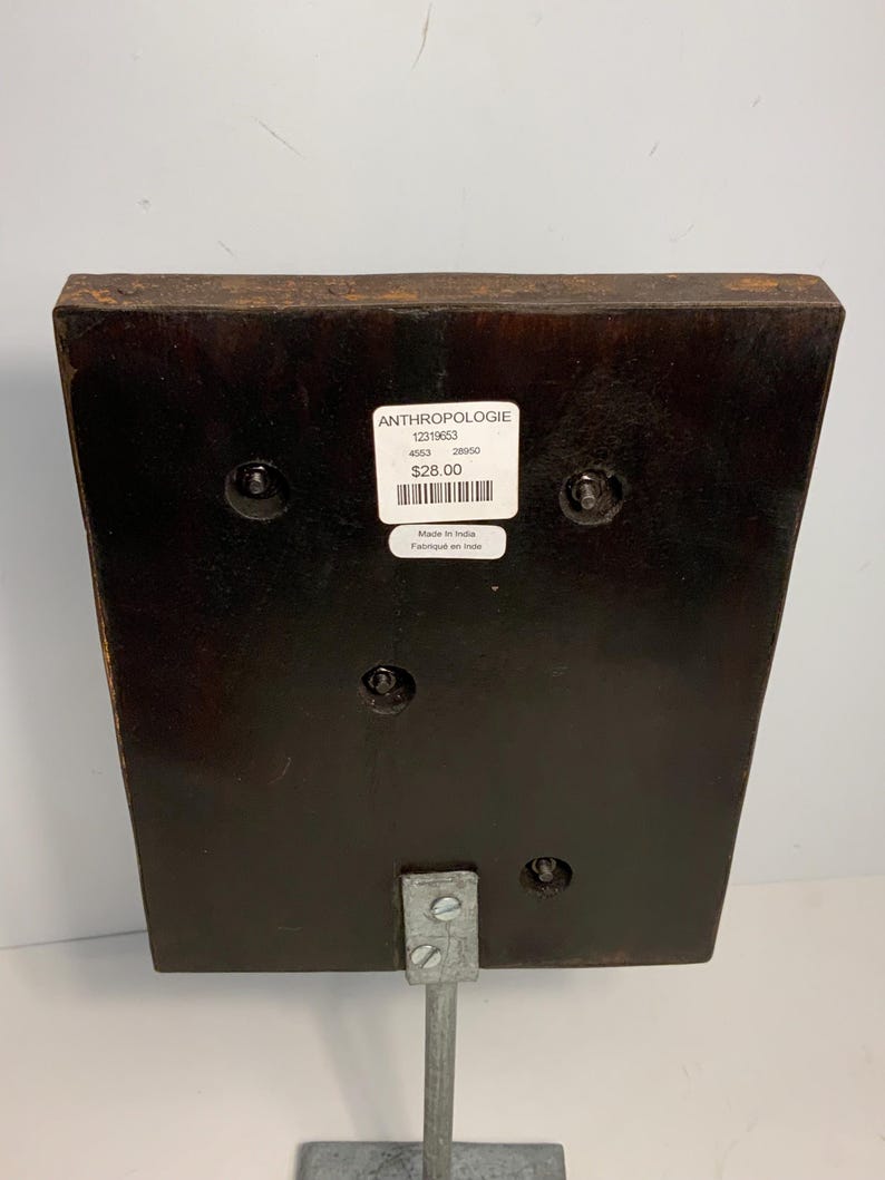 May include: A dark brown wooden decorative piece with a metal stand. The piece has a rectangular shape with rounded edges and is attached to a silver metal stand. A white label with text is visible on the front.