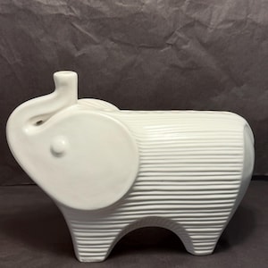May include: A white ceramic elephant figurine featuring a textured body with horizontal ribbed lines. The elephant has a smooth, rounded ear, a raised trunk forming a small opening at the top, and a solid, sturdy base with four short legs.
