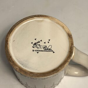 Peut inclure: Mug en c&eacute;ramique cr&egrave;me avec un motif textur&eacute; et une anse incurv&eacute;e. Le fond du mug pr&eacute;sente une illustration noire d'un renne tirant un tra&icirc;neau, avec le texte "by ANTHROPOLOGIE".